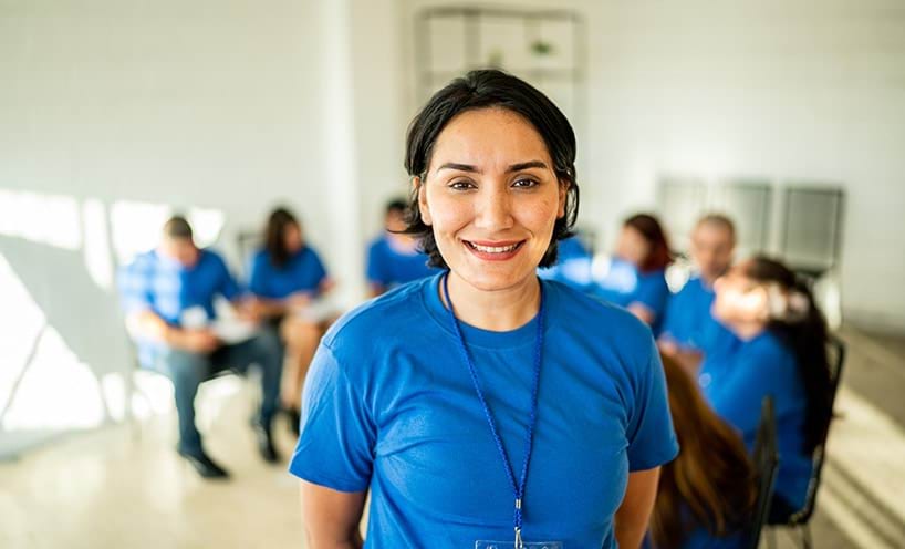 A woman, in a blue t-shirt with a blue lanyard, stands and looks at the camera, while a group of other people sit behind her, also in blue t-shirts.