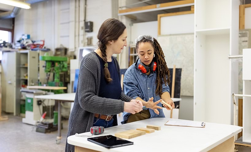 A woman furniture workshop employer standing at table talking and listening to female carpentry apprentice. 