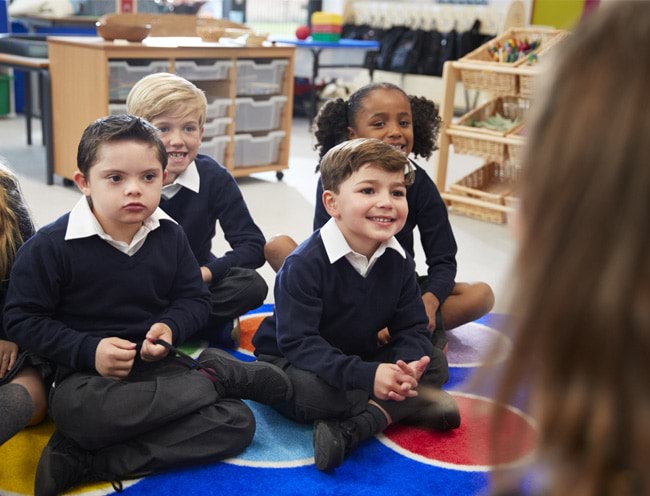 View over female teacher's shoulder of primary school kids sitting in front of her cross legged on the floor of the classroom