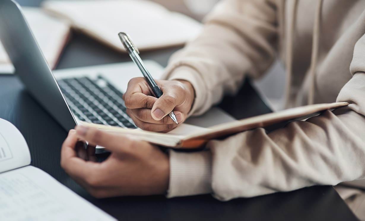 A man takes notes in his notebook, while also using his laptop.